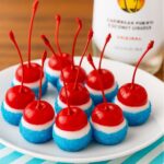 Boozy Cherry Bombs dipped in white chocolate and blue sugar, served on a white plate with a Malibu rum bottle in the background.