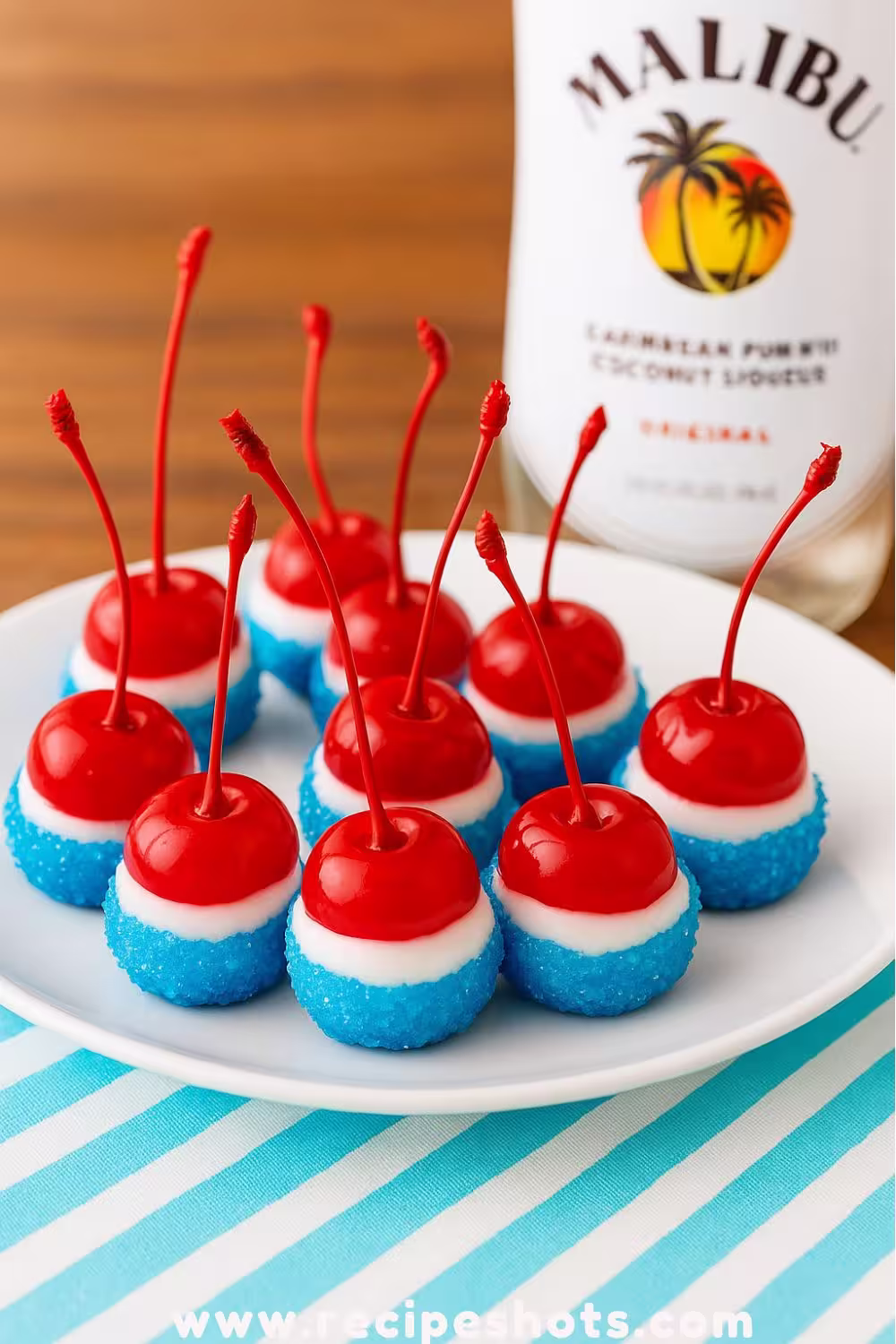 Boozy Cherry Bombs dipped in white chocolate and blue sugar, served on a white plate with a Malibu rum bottle in the background.