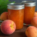 Two jars of homemade vanilla peach jam surrounded by fresh peaches on a rustic wooden surface.