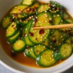 Chopsticks holding sliced cucumber coated in sesame seeds and spicy Asian dressing its Asian Cucumber Salad
