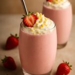 Two glasses of creamy strawberry shortcake protein smoothie topped with whipped cream, crumbled cake, and a fresh strawberry slice.