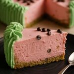 A slice of watermelon cheesecake topped with chocolate chips and surrounded by a green frosting border, served on a black plate.