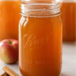 A mason jar filled with golden Apple Pie Moonshine, surrounded by cinnamon sticks and fresh apples.