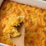 A golden, fluffy serving of pineapple stuffing being scooped from a white baking dish with a wooden spoon.