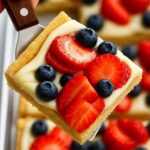 A square slice of fruit pizza topped with fresh strawberries, blueberries, and creamy frosting on a golden cookie crust, held on a spatula.