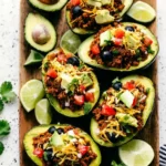 Taco stuffed avocados filled with seasoned ground beef, diced tomatoes, black olives, shredded cheese, avocado chunks, and fresh herbs on a wooden platter.