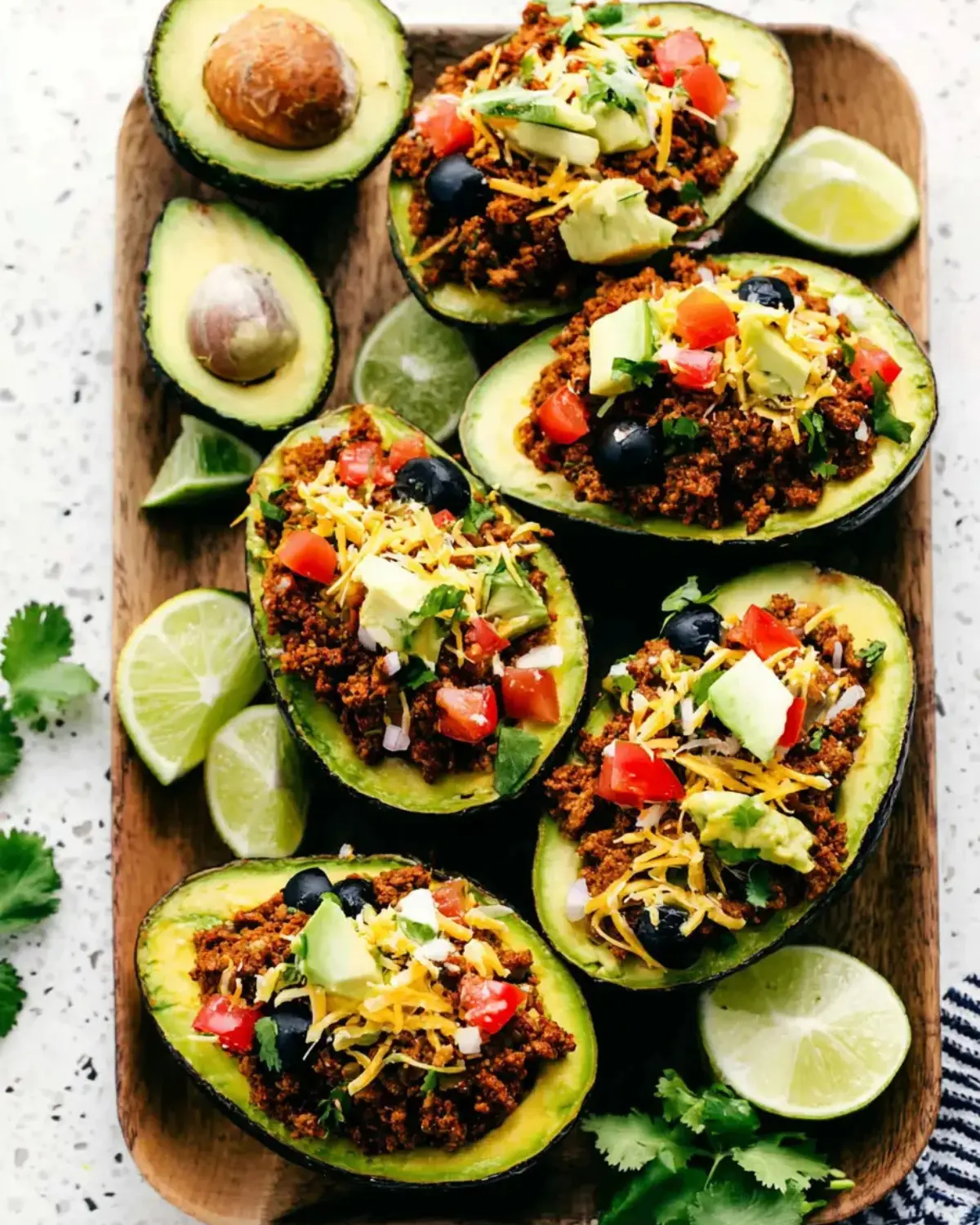Taco stuffed avocados filled with seasoned ground beef, diced tomatoes, black olives, shredded cheese, avocado chunks, and fresh herbs on a wooden platter.