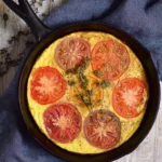 Heirloom tomato frittata cooked in a cast iron skillet, topped with colorful slices of red, yellow, and purple heirloom tomatoes and fresh herbs.
