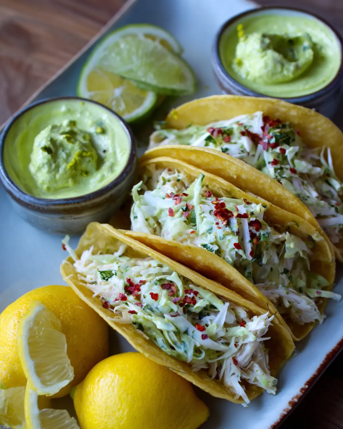 Crab meat tacos recipe served in yellow corn tortillas, filled with creamy crab slaw, garnished with herbs and red pepper flakes, and paired with lemon, lime, and avocado sauce.