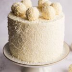 A tall, elegant coconut Raffaello cake covered in white frosting and shredded coconut, topped with white chocolate truffles.