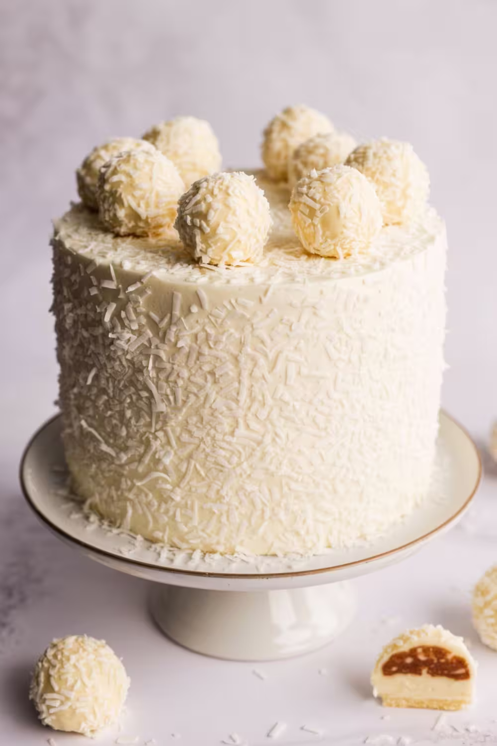 A tall, elegant coconut Raffaello cake covered in white frosting and shredded coconut, topped with white chocolate truffles.
