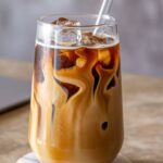 A refreshing glass of iced caramel coffee with swirls of cream and ice cubes, served with a glass straw.
