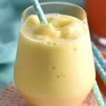 A glass of creamy yellow Creamsicle Slushy with a blue polka-dot straw on an orange plate. Title: Refreshing and Creamy Creamsicle Slushy Drink