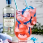 A vibrant Shark Attack Punch Cocktail garnished with gummy sharks in a tall glass filled with ice, with a bottle of vodka in the background.