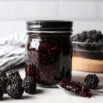 A glass jar filled with homemade black raspberry jam, surrounded by fresh black raspberries and a spoonful of jam on a white surface.