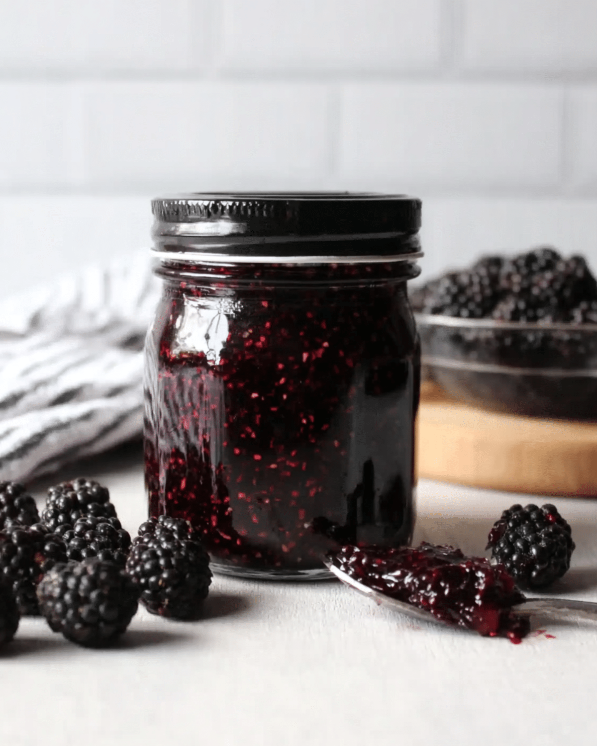 A glass jar filled with homemade black raspberry jam, surrounded by fresh black raspberries and a spoonful of jam on a white surface.