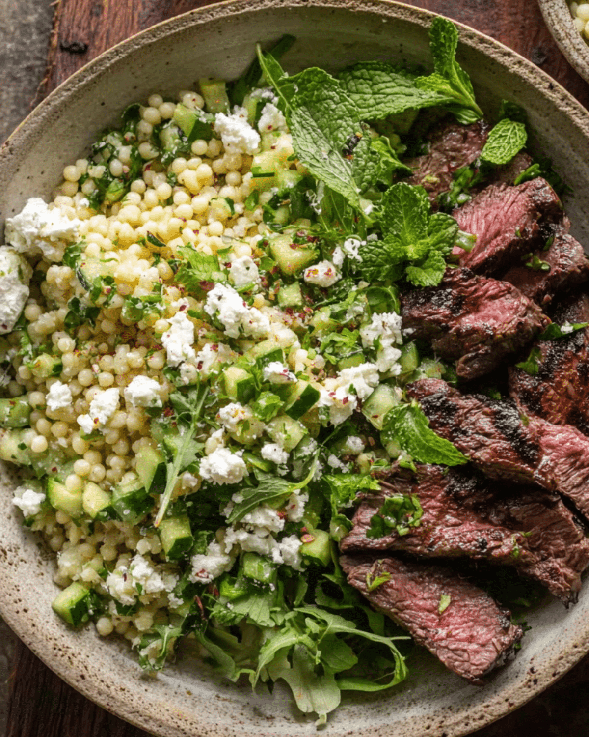 Grilled skirt steak served with lemon herb couscous salad, fresh mint, cucumbers, feta cheese, and arugula in a rustic ceramic bowl.