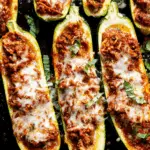 Ground beef zucchini boats topped with melted cheese and fresh herbs on a dark baking tray.