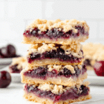 A stack of sour cherry crumble bars with buttery crust, juicy cherry filling, and crumbly topping on a white surface.