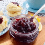 A glass jar of sour cherry jam with a spoon inside, served alongside scones on a wooden board.