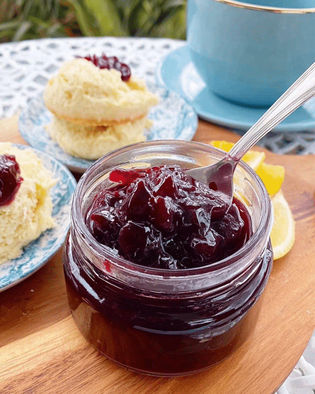 A glass jar of sour cherry jam with a spoon inside, served alongside scones on a wooden board.