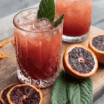 A refreshing glass of blood orange and mulberry leaf solid drink with ice, garnished with fresh mulberry leaves and surrounded by sliced blood oranges.