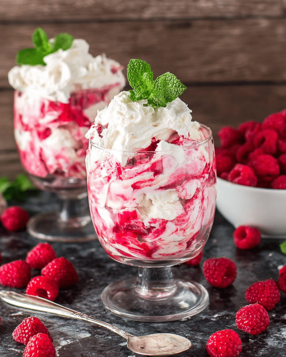 Glasses filled with easy raspberry eton mess made with whipped cream, raspberry sauce, and meringue, garnished with mint.
