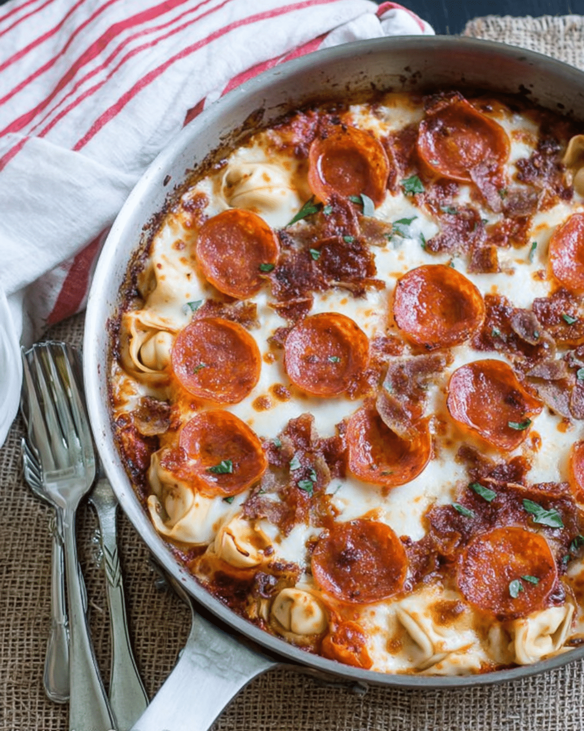 A skillet of baked meat lovers pizza tortellini topped with melted cheese, pepperoni, bacon, and herbs, with cheese tortellini peeking through.