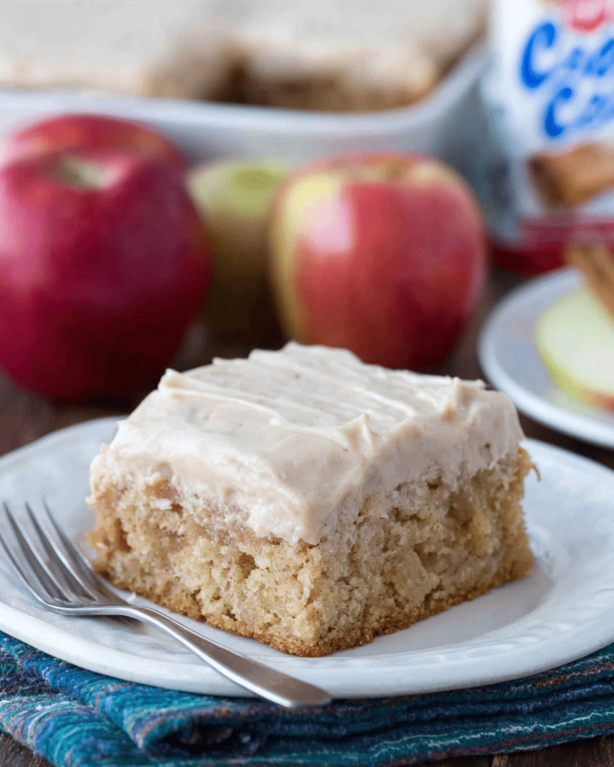 Slice of apple cinnamon sheet cake topped with creamy frosting, served on a white plate with a fork, and fresh apples in the background.