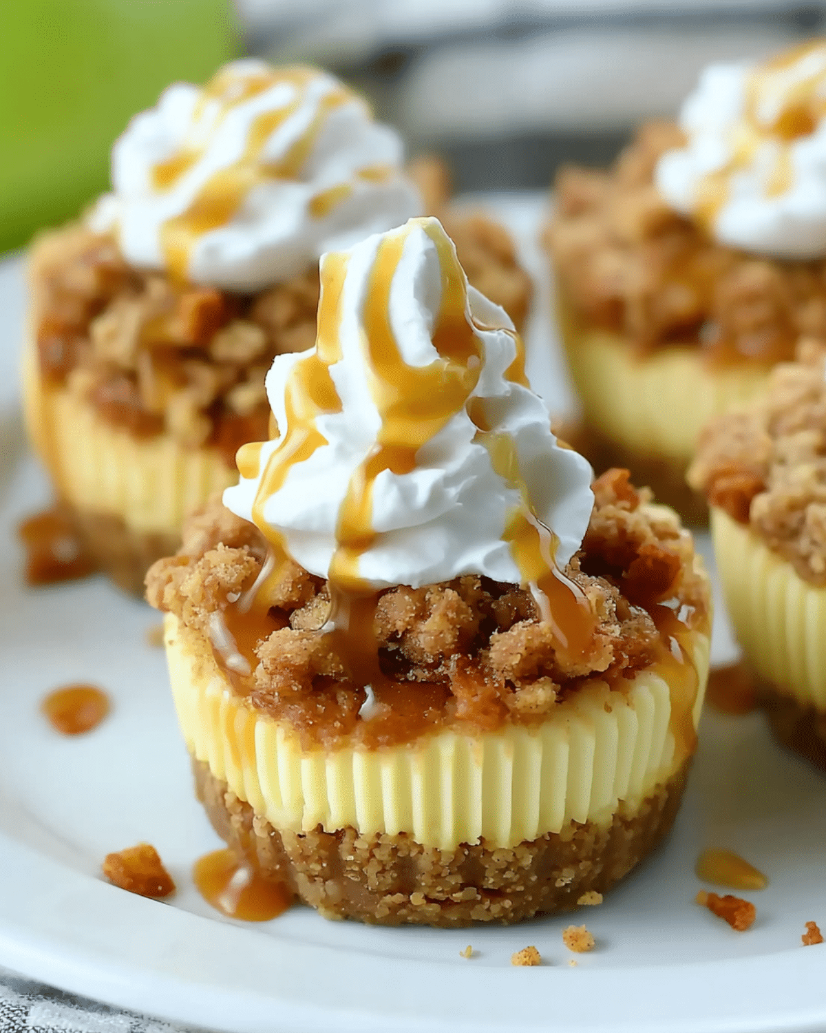 Apple crisp mini cheesecake topped with whipped cream and caramel drizzle, set against a background of green apples.