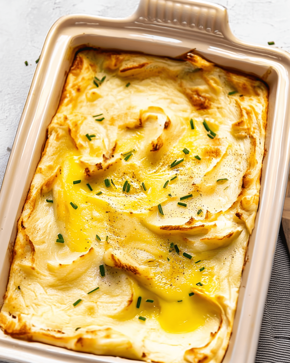 Golden-topped Creamiest Baked Mashed Potatoes in a ceramic baking dish, garnished with melted butter and chopped chives.