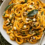 A bowl of creamy pumpkin pasta garnished with fresh herbs, chopped nuts, and sage leaves.