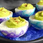 Creepy deviled eggs for Halloween with vibrant purple and blue marbled whites and green filling, topped with red seasoning.