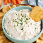 Creamy French onion dip garnished with chopped parsley in a blue bowl, surrounded by potato chips with one chip dipped in.
