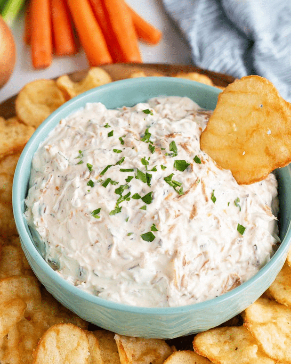 Creamy French onion dip garnished with chopped parsley in a blue bowl, surrounded by potato chips with one chip dipped in.