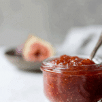 A glass jar filled with homemade fig jam, with a spoon inside and fresh figs blurred in the background.