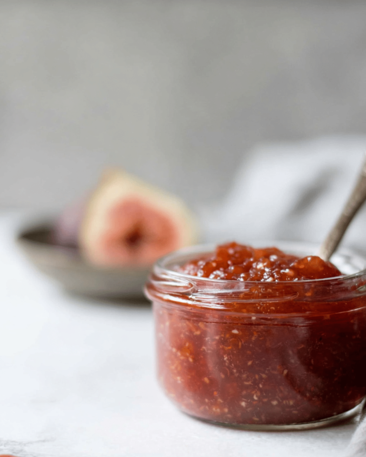 A glass jar filled with homemade fig jam, with a spoon inside and fresh figs blurred in the background.