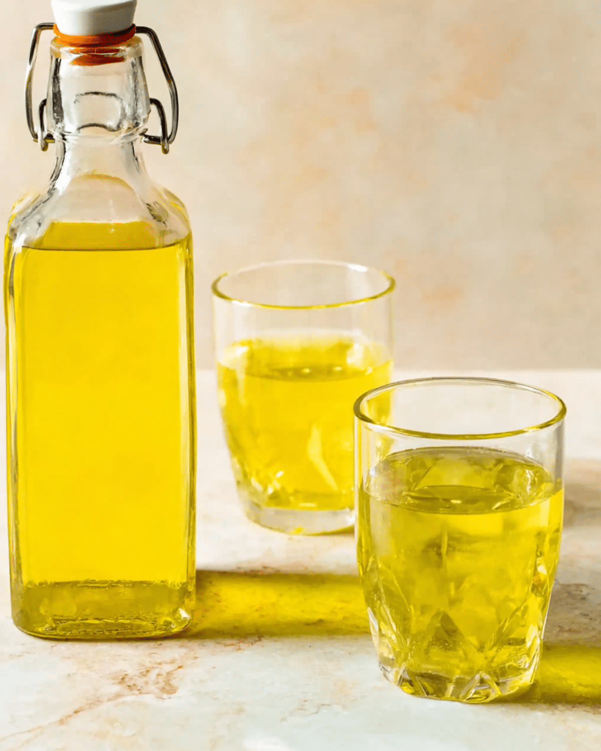 Bottle and glasses filled with bright yellow Limoncello on a light marble surface.