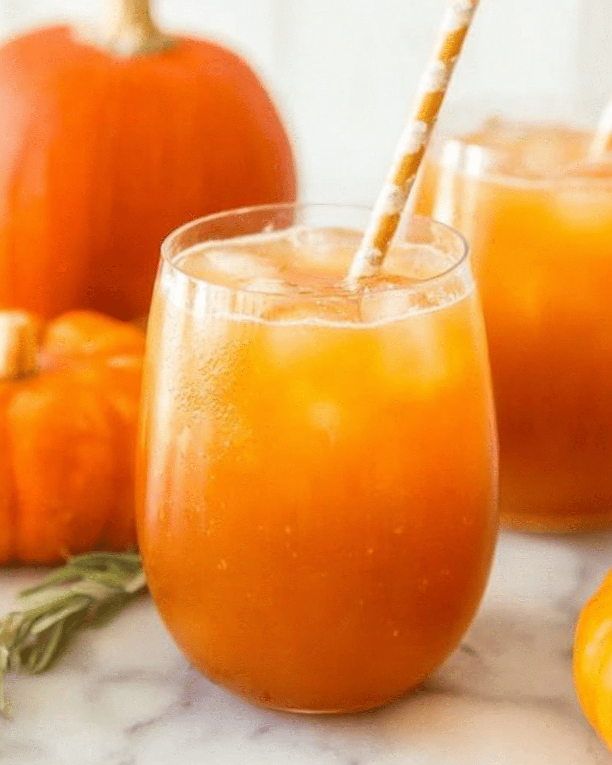 A chilled glass of magical Harry Potter pumpkin juice with ice and a striped straw, surrounded by fresh pumpkins.