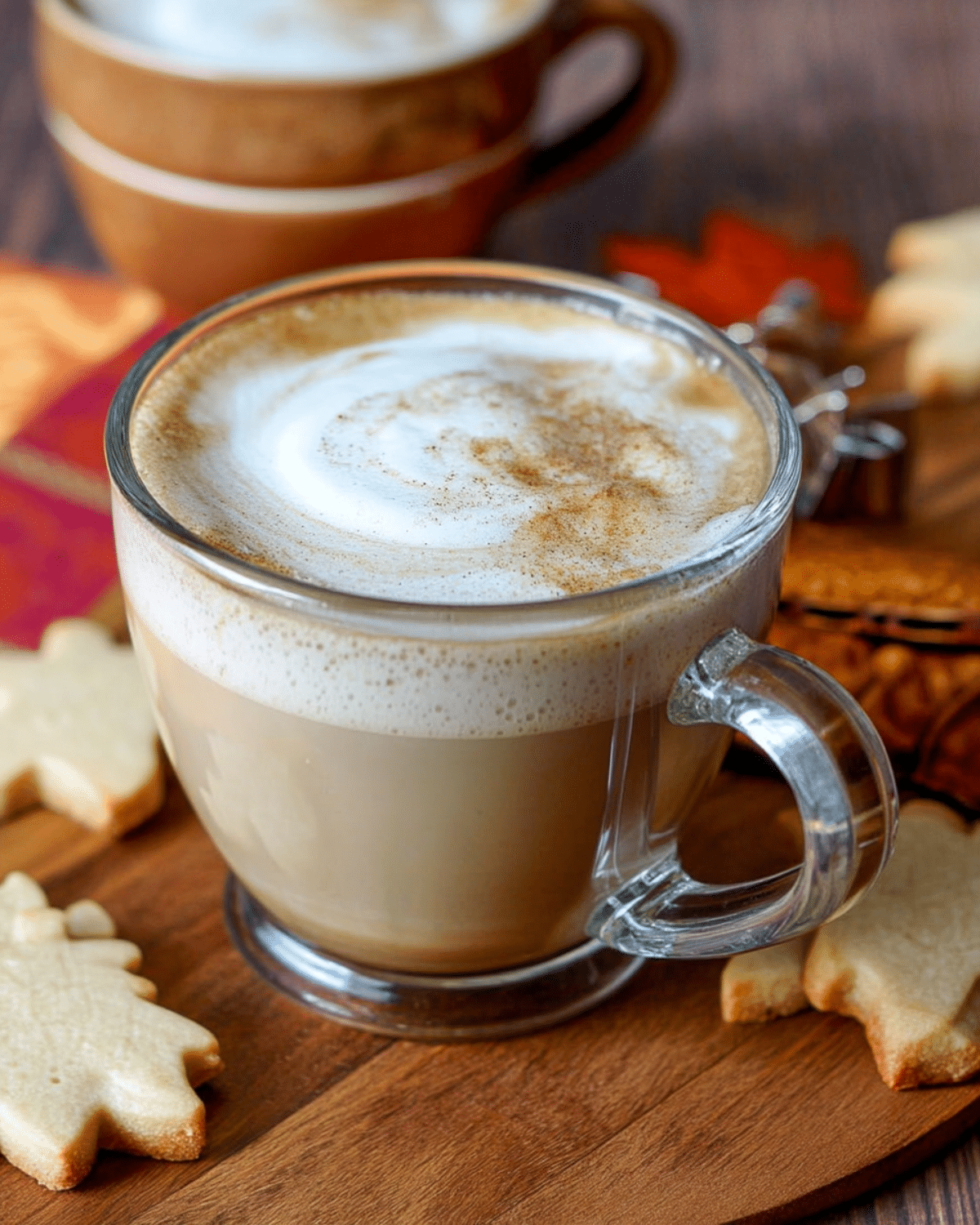 Glass mug filled with maple cinnamon latte topped with frothy milk and cinnamon, surrounded by leaf-shaped cookies on a wooden board.