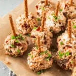 Mini cheese ball bites coated in chopped bacon and herbs, each with a pretzel stick, arranged on parchment paper.