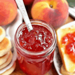 Jar of nectarine jam without pectin with a spoon inside, surrounded by fresh nectarines, toast, and biscuits.