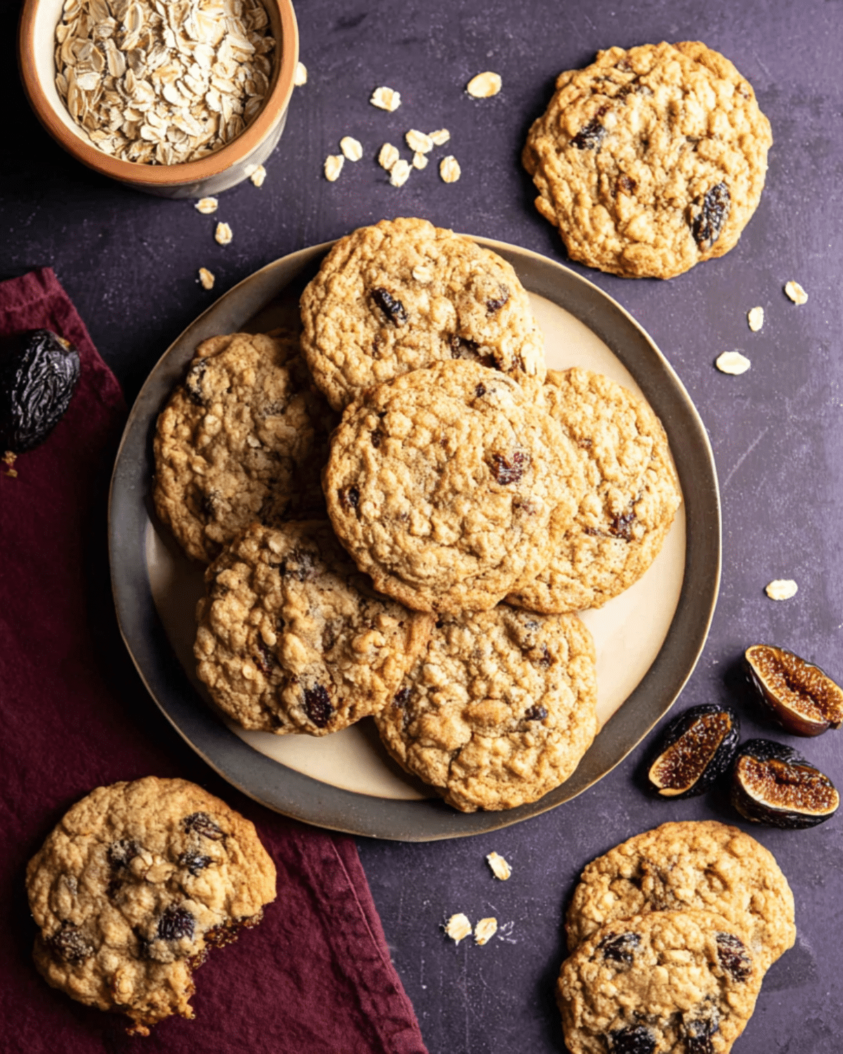 A plate of oatmeal fig cookies surrounded by dried figs, oats, and additional cookies on a dark surface.