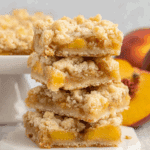 Stack of peach bars with crumbly topping and peach filling, placed on a white marble coaster with fresh peaches in the background.