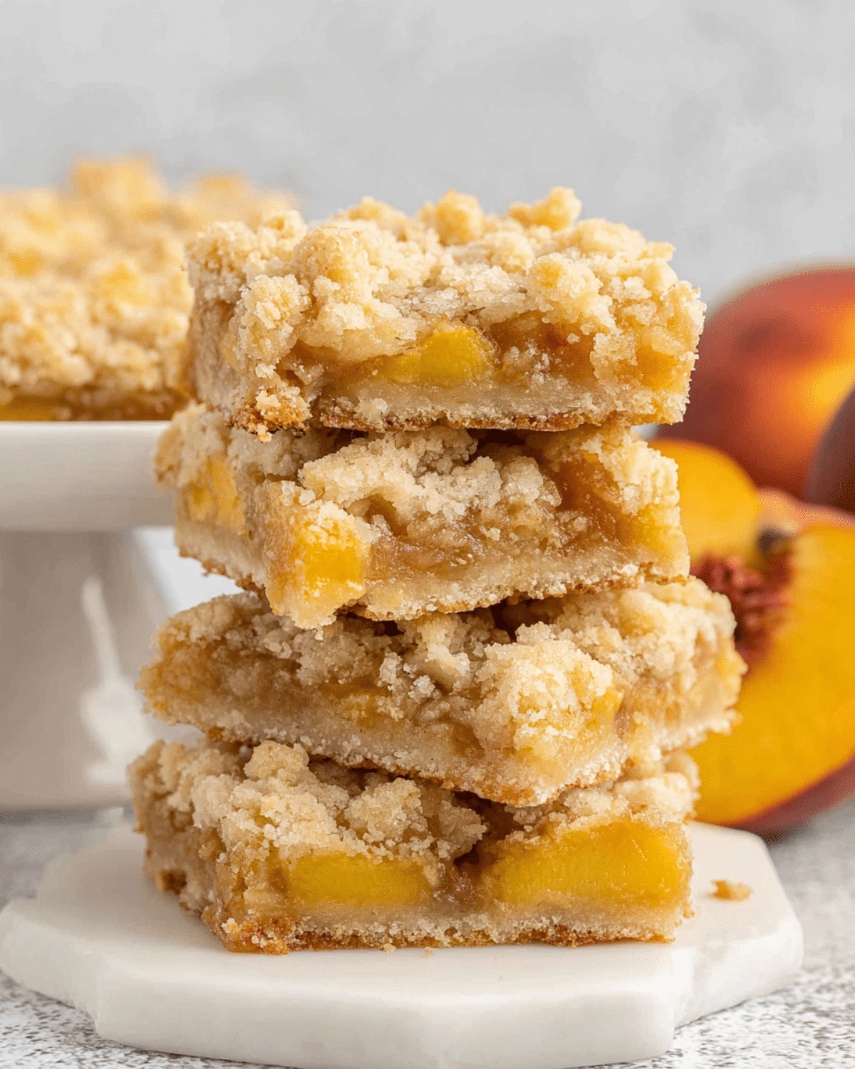 Stack of peach bars with crumbly topping and peach filling, placed on a white marble coaster with fresh peaches in the background.