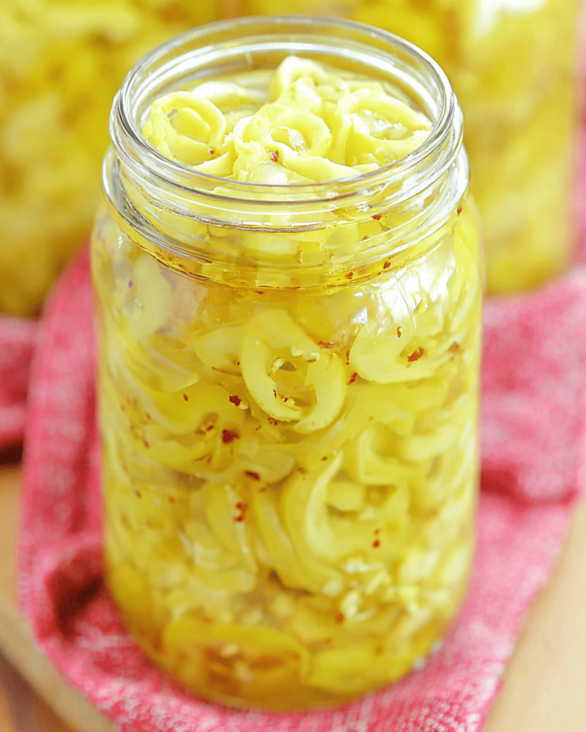 Jar of pickled banana peppers in brine with red pepper flakes, set on a red cloth.