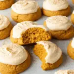 Soft pumpkin cookies topped with cream cheese frosting and sprinkled with cinnamon, with one cookie broken in half to reveal its fluffy texture.