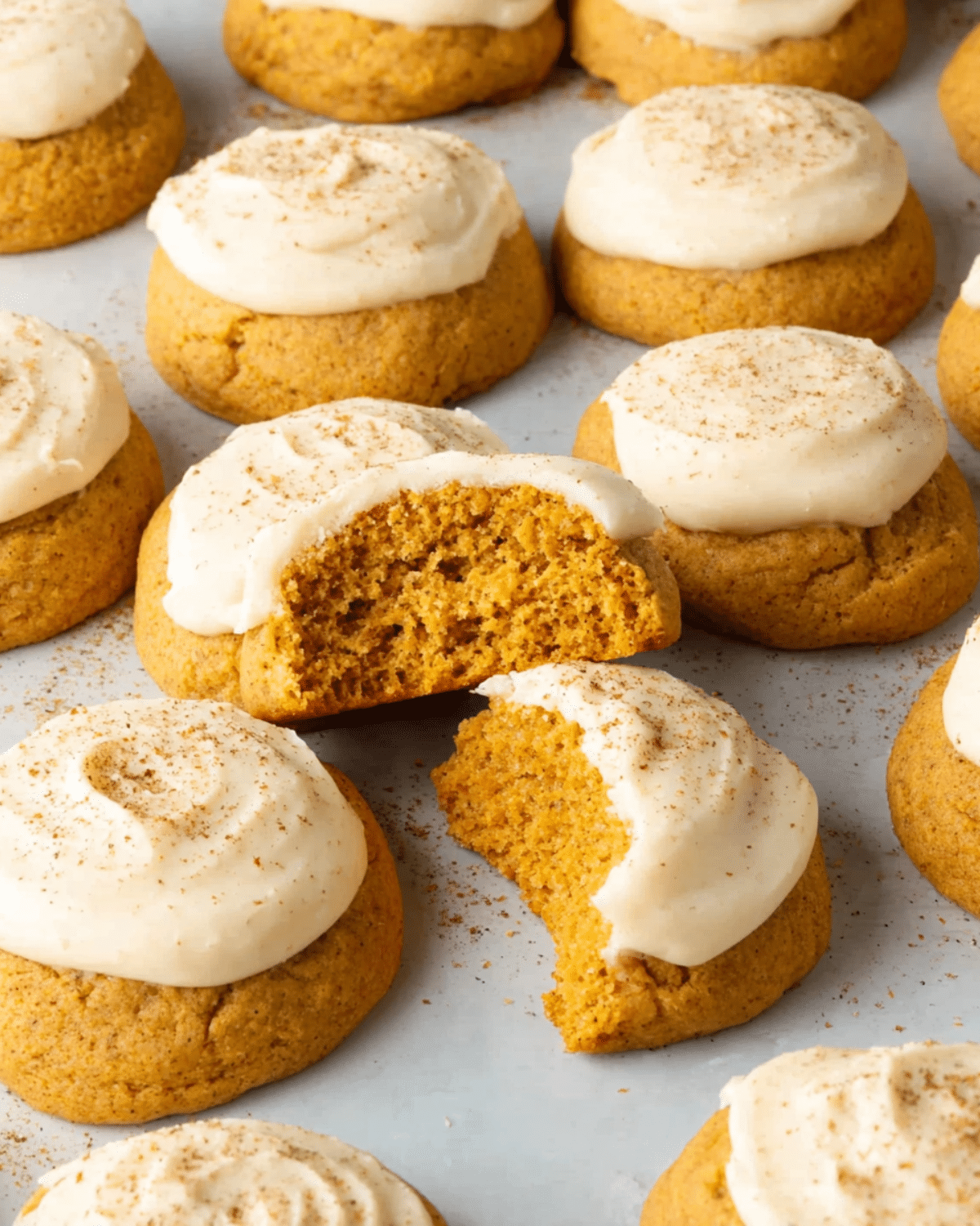 Soft pumpkin cookies topped with cream cheese frosting and sprinkled with cinnamon, with one cookie broken in half to reveal its fluffy texture.