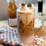 Glass of toasted marshmallow iced coffee topped with two charred marshmallows, set on a checkered towel with coffee beans and mini marshmallows around.