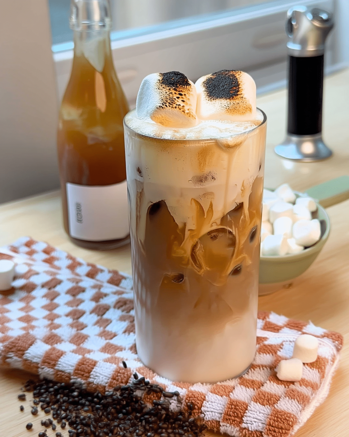Glass of toasted marshmallow iced coffee topped with two charred marshmallows, set on a checkered towel with coffee beans and mini marshmallows around.